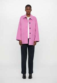 Jachetă oversized dintr-un amestec de lână roz, cu guler, butoane frontale și două buzunare, purtată peste o cămașă albă și asortată cu pantaloni în dungi închise.