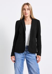 Černý přiléhavý blazer s texturovanou tkaninou, dvěma předními kapsami a klopovým límcem, spárovaný se světle modrými džíny a bílým topem.