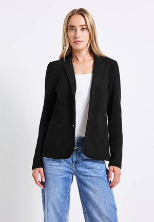 Blazer noir ajusté en tissu texturé, avec deux poches avant et un col à revers, associé à un jean bleu clair et un haut blanc.