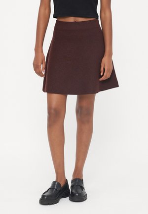 Jupe a-ligne en tricot marron avec une taille ajustée, descendant au-dessus du genou. Associée à des chaussures en cuir noires avec une semelle épaisse.