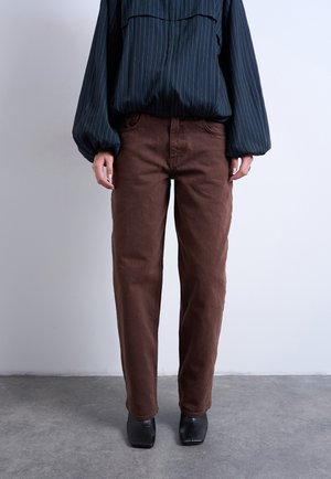 Persoon draagt bruine rechte broekspijpen, zwarte schoenen met vierkante neus en een donkere blouse met lange mouwen en fijne strepen, tegen een effen muur.