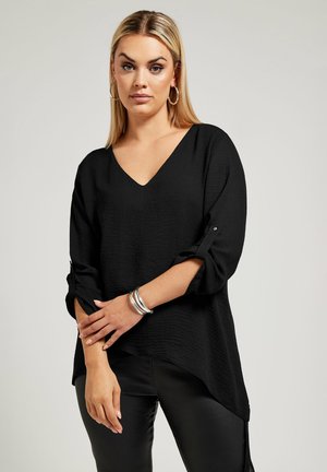 Zwarte v-hals top met opgerolde mouwen, gestructureerde stof, asymmetrische zoom, gecombineerd met zilveren armband en zwarte broek.