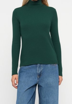 Woman wearing a dark green turtleneck sweater and light blue high-waisted jeans standing against a white background.