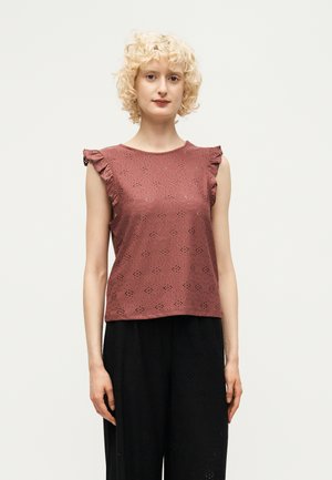 Giovane donna con capelli biondi corti e ricci che indossa un top smanicato malva con occhielli e dettagli di volant e pantaloni neri a gamba larga.