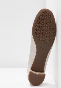 Chaussure beige avec une tige synthétique lisse, un bout arrondi et une semelle en caoutchouc marron comportant un motif texturé et perforé. Marque Geox visible.