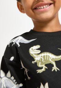 Svart långärmad skjorta med dinosaurieskeletttryck i vitt och olivgrönt. Rund halsringning och mjuk bomullstextur.