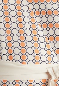 Tissu avec un motif répété de points orange à l'intérieur de cercles noirs sur fond blanc, muni d'une ceinture blanche nouée.