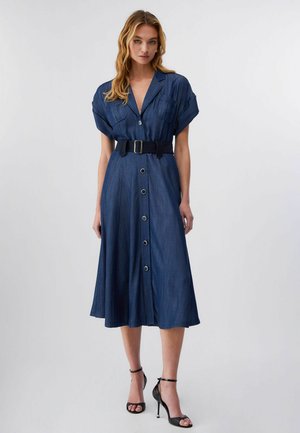 Donna che indossa un vestito blu navy a metà lunghezza con bottoni sul davanti, maniche corte, tasche frontali, cintura nera e sandali neri con tacco alto.