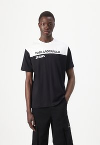 Fekete-fehér rövid ujjú póló, amelyen a "KARL LAGERFELD JEANS" felirat található, pamutból készült, sima textúrával és kerek nyakú kialakítással.