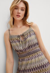 Bunt gekleidetes Kleid mit einem gerafften Oberteil, das ein Bindedetail am Ausschnitt aufweist. Das Muster umfasst florale und geometrische Designs in zarten Pastellfarben.