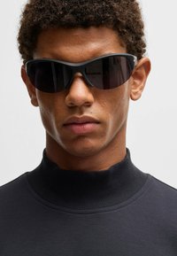 Svarta solglasögon med en smidig, omslutande design och mörka linser, kombinerat med en svart tröja med hög krage och ribbad textur.