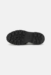 Semelle de chaussure noire avec des crampons profonds et robustes et des motifs en forme de croix, conçue pour assurer l’adhérence sur des surfaces rugueuses.