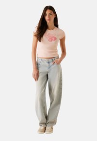 Lichtroze fitted t-shirt met "Guess" grafiek en aardbeienontwerp, gecombineerd met lichtblauwe baggy jeans en beige sneakers.