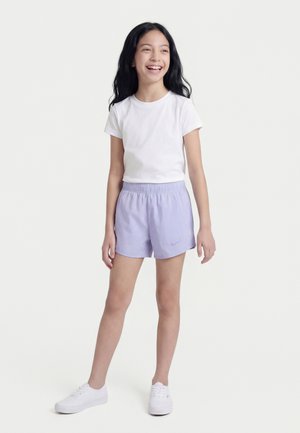 Fille debout et souriante portant un t-shirt blanc, un short Nike violet clair et des baskets blanches sur un fond uni.