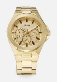 Gouden metalen horloge met een gestructureerde wijzerplaat, drie sub-dials en gedurfde uurmarkers. Voorzien van een chunky armband en een kroon aan de zijkant.