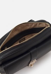 Černá texturovaná kožená taška se zlatým zipem a sponou; interiér má béžovou podšívku a vzorovaný branding.