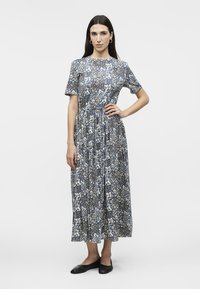Femme debout, main sur la hanche, portant une robe midi bleue à motifs floraux et des ballerines noires sur un fond blanc uni.