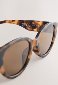 Kilpikonnakuosisunglasses, joissa on harmaa kehys, ruskeat linssit ja pyöristetty kissansilmämuoto. Sivussa on brändin ja vaatimustenmukaisuuden merkinnät.