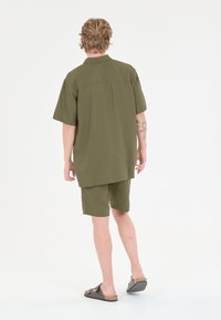 Oliivinvihreä rennosti istuva lyhythihainen paita, jossa on takasauma, yhdistettynä samansävyisiin shortseihin. Mallilla on päällään ruskeat sandaalit.