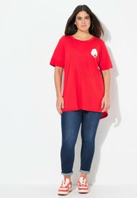 T-shirt en coton rouge avec des manches courtes et un graphique circulaire blanc sur la poitrine, associé à un jean bleu foncé et des baskets rouges.