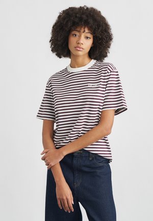 Giovane donna con capelli ricci che indossa una t-shirt a righe bordeaux e bianche e jeans blu scuro, in piedi con le braccia incrociate.