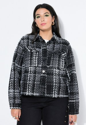 Ung kvinde med langt mørkt hår iført gyldne hoops-øreringe, grøn øjenskygge, en sort-hvid ternet jakke og sorte bukser med gyldne knapper.