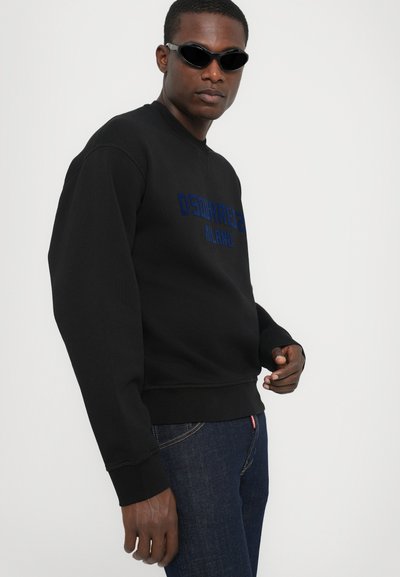 Sweat-shirt noir avec le texte bleu "DSquared2 MILANO", encolure ronde et poignets côtelés. Associé à un jean en denim foncé et des lunettes de soleil.