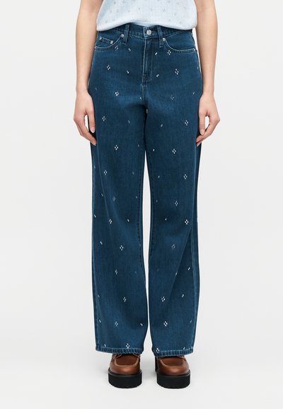 Γυναίκα που φοράει σκούρα μπλε τζιν παντελόνι με ψηλή μέση και φαρδιά μπατζάκια, διακοσμημένο με μικρές λευκές κεντημένες τελείες, συνδυασμένο με καφέ παπούτσια πλατφόρμας.