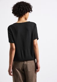 Top corto nero con texture e vestibilità ampia, abbinato a pantaloni marrone chiaro. La vista posteriore mostra un collo semplice e un tessuto fluido.