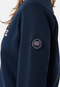 Marineblauwe sweatshirt gemaakt van zacht materiaal, met een ronde geborduurde patch op de linker mouw met "Seaside" en "Established 1976."