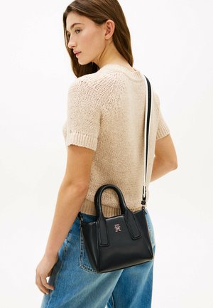 Vrouw in beige gebreide korte mouw trui en blauwe spijkerbroek met een zwarte leren handtas met lange en korte hengsels over de schouder gedragen.