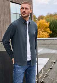 Mörkblå skjorta med knäppning över en vit T-shirt, tillsammans med blå jeans. Skjortan har logotypdetaljer och långa ärmar.