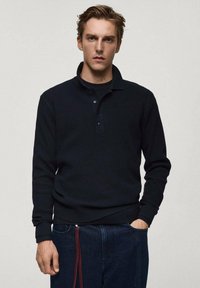 Black ribbed long-sleeve polo shirt with a collar, featuring a three-button placket. Paired with dark blue denim pants and a decorative zipper pull.