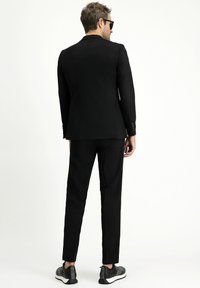 Schwarzer Anzug mit einem taillierten Sakko und schmal zulaufenden Hosen, aus glattem Stoff. Kombiniert mit schwarzen Sneakers, von hinten betrachtet.