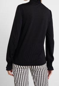 Schwarzes Langarmtop mit rüschigem Ausschnitt, glatter Textur und gerafften Manschetten. Kombiniert mit schwarz-weißen Hosen mit vertikalen Streifen.