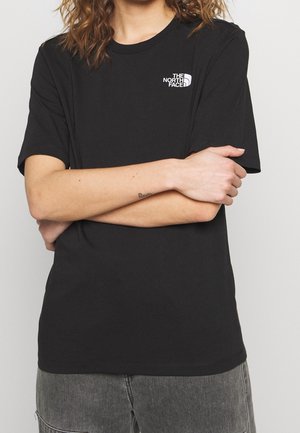Osoba o kręconych włosach, ubrana w czarną koszulkę The North Face z krótkim rękawem i szare dżinsowe szorty, z rękami skrzyżowanymi na wysokości talii.