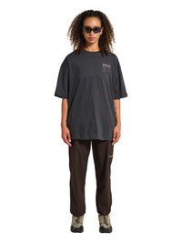 Kobieta z kręconymi włosami, nosząca czarne okulary przeciwsłoneczne, oversize ciemnoszarą koszulkę, brązowe spodnie i beżowe buty trekkingowe, stojąca na białym tle.