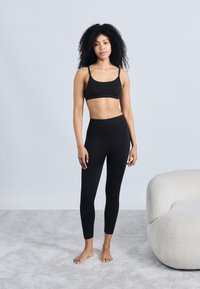 Schwarzer Sport-BH und hoch taillierte Leggings aus dehnbarem Material mit glatter Textur und enganliegendem Schnitt. Im Hintergrund weicher grauer Teppich und ein gemütlicher Sessel.