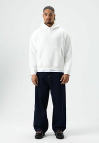 Witte hoodie met capuchon en een zak aan de voorkant, gecombineerd met donkerblauwe wijde jeans en zwarte schoenen met roze veters, tegen een effen achtergrond.