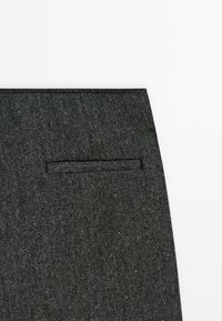 Harmaa herringbone-kangas, jossa on kapea sovitus, litteä tasku ja hillitty rakenne, korostaa kudoksen sisällä olevaa monimutkaista kuviointia.