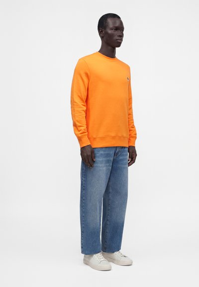 Homme debout portant un sweat-shirt orange vif à manches longues, un jean bleu ample et des baskets blanches sur un fond clair uni.