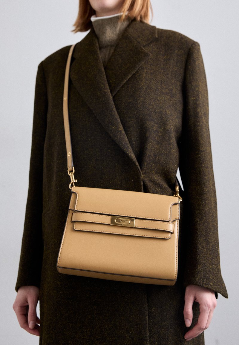 Borsa a mano in pelle tan con design strutturato, dotata di una chiusura in metallo dorato e di una tracolla rimovibile, tenuta sotto un cappotto marrone scuro.