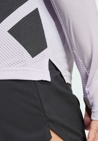 Haut de sport à manches longues en violet clair avec des panneaux en maille gris foncé. Présente une fente sur le côté pour faciliter les mouvements, associé à un short noir.
