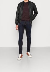 Svart läderbomberjacka över en vinröd och marinrandig skjorta, kombinerad med mörkblå slim-fit jeans och bruntonerade kanvas sneakers.