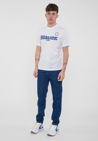 T-shirt bianca con logo blu "Blauer USA", abbinata a jeans in denim blu scuro e sneakers bianche con accenti blu e neri.