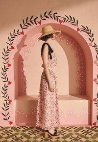 Kvinna i en blommig maxiklänning och halmhatt står i profil mot en rosa valvvägg med röda hjärt- och lövmönster.