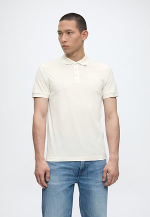 Muž oblečený v jednofarebnom bielom polotriku s krátkym rukávom a svetlomodrých džínsoch, stojaci pred jednofarebným bielym pozadím a pozerajúci sa do strany.