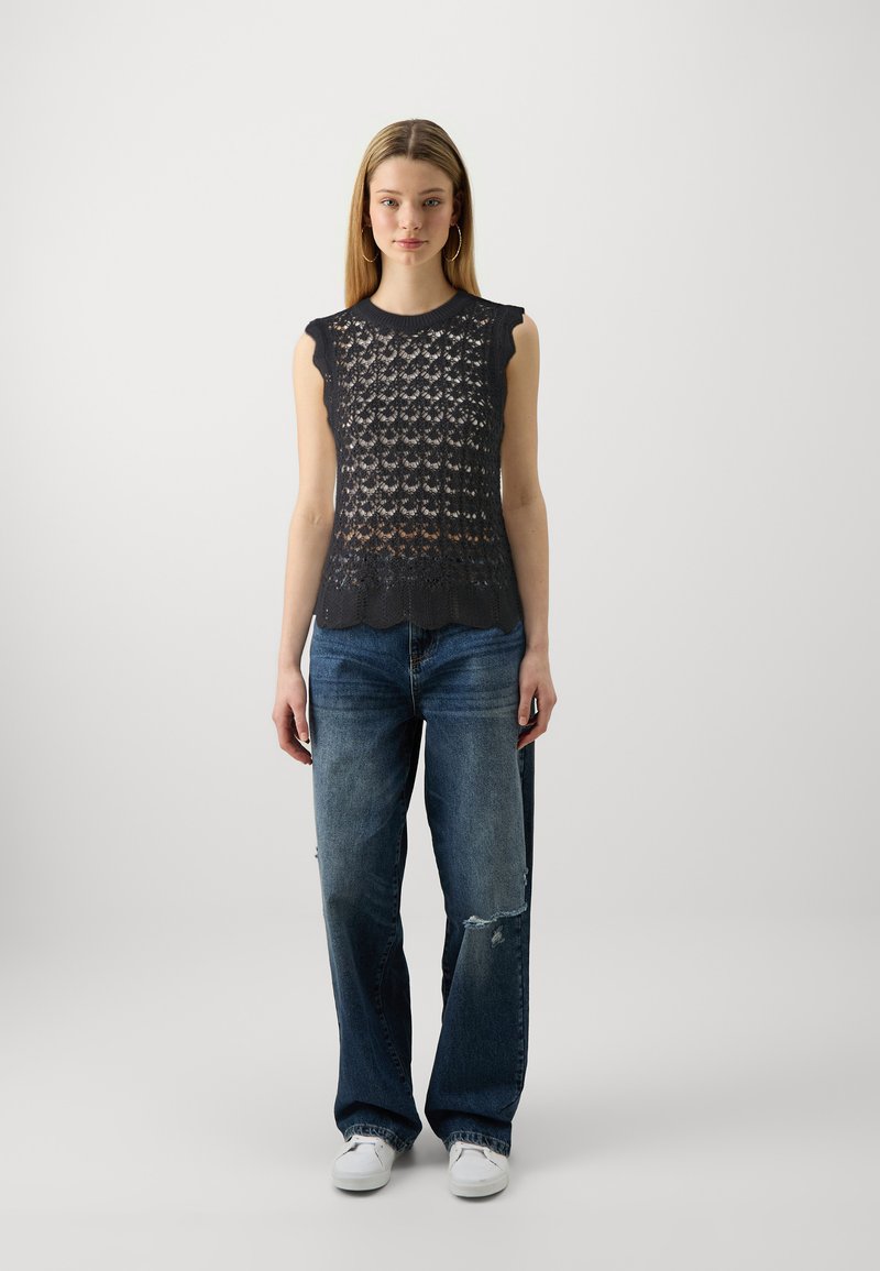 Ärmelloses Top aus Spitzenstrick in Dunkelgrau, mit gewellten Kanten und lässiger Passform, kombiniert mit ausgewaschenen blauen weiten Jeans und weißen Sneakern.