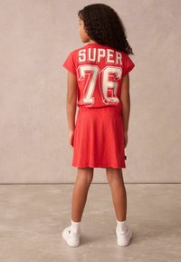 Röd klänning med en åtsittande topp som har "SUPER 76" i vit lettering. Har korta ärmar, en utsvängd kjol och vita sneakers.