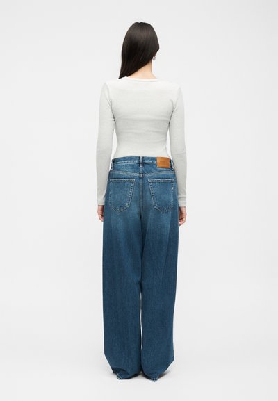 Γυναίκα με μακριά σκούρα μαλλιά, φορώντας στενό λευκό μακρυμάνικο τοπ και φαρδιά μπλε τζιν παντελόνια, στέκεται με την πλάτη.